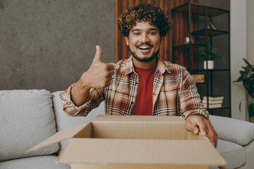 Young smiling happy Indian man wears casual clothes hold cardboard box unpacking show thumb up sits on grey sofa couch stay at home hotel flat rest relax spend free spare time in living room indoor.