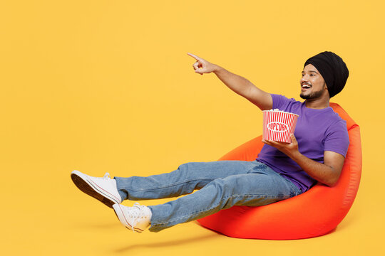 Full body devotee Sikh Indian man ties his traditional turban dastar wear purple t-shirt sit in bag chair hold bucket of popcorn watch movie film isolated on plain yellow background studio portrait.