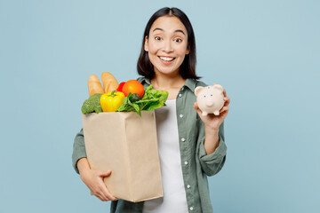 Fototapeta na wymiar Young surprised happy woman wear casual clothes hold brown paper bag with food products pig money box isolated on plain blue cyan background studio portrait. Delivery service from shop or restaurant.