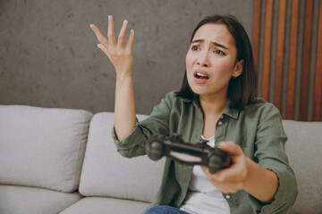 Young sad woman of Asian ethnicity wears casual clothes hold play pc game with joystick console sit on grey sofa couch stay at home hotel flat rest relax spend free spare time in living room indoor.