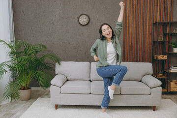 Full body young smiling fun cool woman of Asian ethnicity wears casual clothes dancing stand near grey sofa dance couch stay at home hotel flat rest relax spend free spare time in living room indoor.
