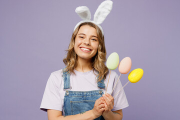 Young cheerful woman wear casual clothes bunny rabbit ears hold in hands palms colorful eggs on sticks isolated on plain pastel light purple background studio portrait. Lifestyle Happy Easter concept.