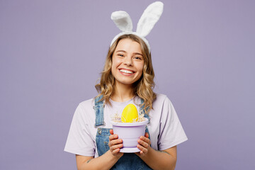 Young smiling cool woman wearing casual clothes bunny rabbit ears hold in hands big yellow egg in pot isolated on plain pastel light purple background studio portrait. Lifestyle Happy Easter concept.