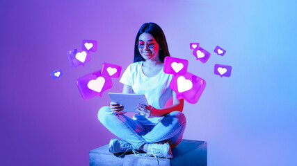 Smiling young asian woman in glasses typing on tablet, has romantic chat with hearts on neon background