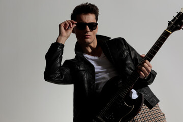 sexy young man in black leather jacket fixing glasses and playing guitar
