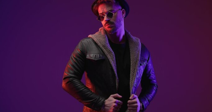 confident cool man puling leather jacket and wool cardigan, adjusting hat and looking and moving to side in front of grey background with colorful lights