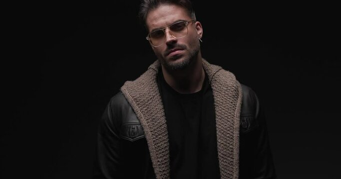 project video with flash lights of cool young man wearing black leather jacket, glasses and wool cardigan looking to side, touching and fixing beard on black background