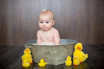 Cute baby playing with rubber duck while sitting in metal basin - obrazy, fototapety, plakaty