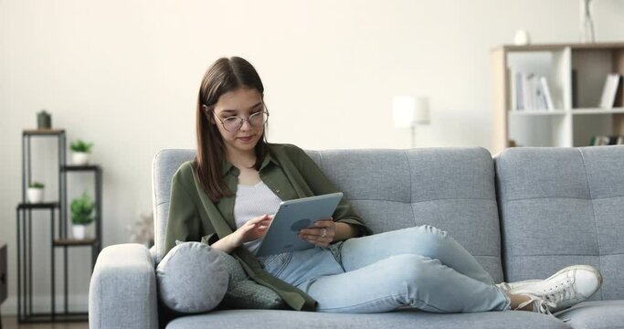 Relaxed 16s teen girl in glasses spend time with digital tablet, relax on couch in living room, smile, enjoy remote chat with friends, use app, watch videos, share messages via app. Leisure, internet