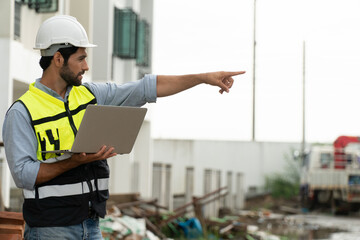 Foreman with Yellow helmet holding laptop in constuction site pointing finger to building