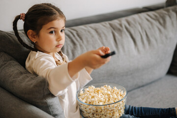 Cute little girl using remote controller of TV and watching cartoons. Four year girl eating popcorn on sofa