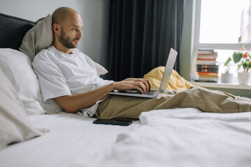 A man works on a laptop in bed in a bedroom. Freelancer at home. A programmer or developer works remotely.