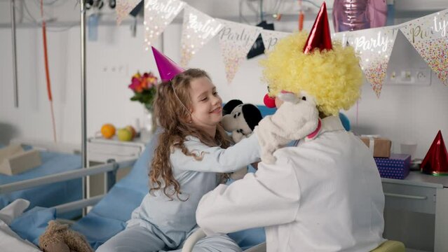 Happy doctor with clown red noses celebrating birthday with little girl in hospital room.