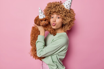 Horizontal shot of curly haired young woman holds small adorable poodle puppy near face keeps lips rounded wear party hats isolated over pink background celebrate birthday of domestic animal.