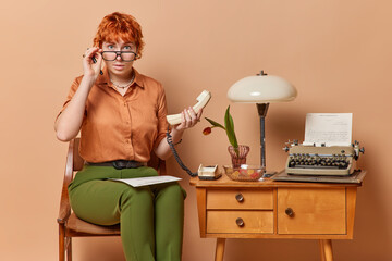 Horizontal shot of amazed astonished female teacher keeps hands on rim of spectacles holds handset poses on chair with papers on knees makes article on typewriter isolated over brown background