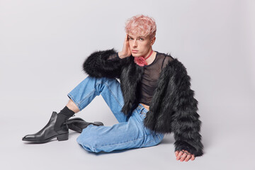 Full length horizontal shot of serious pink haired gay dressed in black fur coat jeans and leather shoes has non traditional orientation isolated over white studio background. Sexy glamour trans man - obrazy, fototapety, plakaty