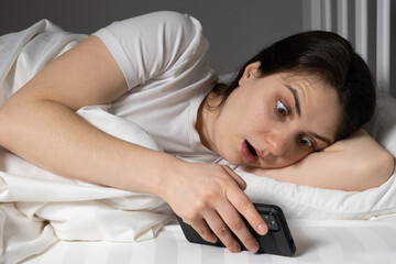 A young woman looks in surprise with her mouth open at the screen of a smartphone lying in bed, a gadget before going to bed.