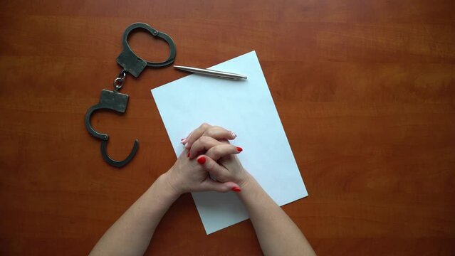 Women's manicured hands are nervously twirling a pen, next to handcuffs and a blank sheet of paper