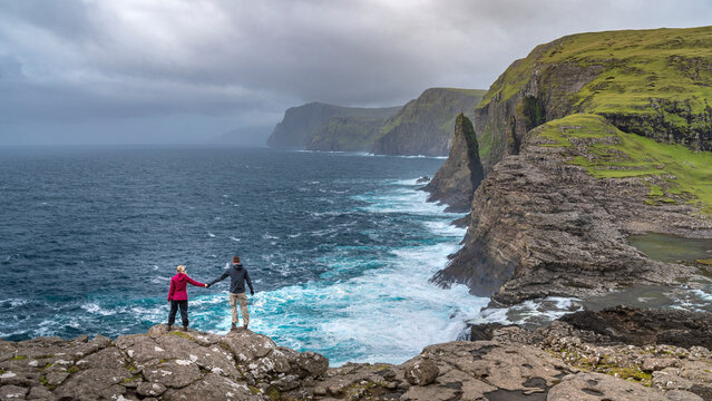 Zwei Wanderer stehen Hand in Hand am B&oslash;sdalafossur Wasserfall und blicken auf den Geituskorardrangur Felsen. Das Meer brandet gegen die Klippen.