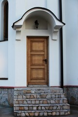 Fototapeta na wymiar A light brown wooden door with a black lock, above the door is a lantern, the building is white, the lower part is decorated with stones and stairs