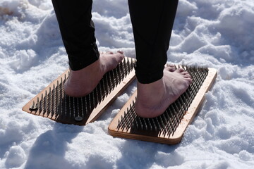 A woman practicing yoga therapy stands on a board with nails against a background of snow.