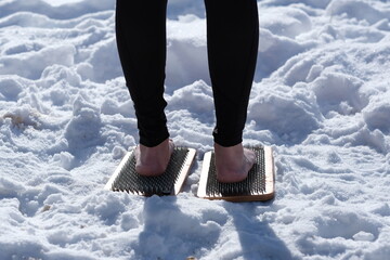 A woman practicing yoga therapy stands on a board with nails against a background of snow.
