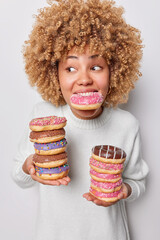 Vertical shot of curly haired woman poses with stack of appetizing glazed doughnuts breaks diet...
