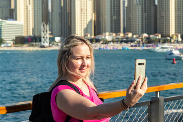 a photo of a blonde tourist girl with a backpack standing on a Dubai embankment with an incredible view of the sea and the nearby modern skyscrapers