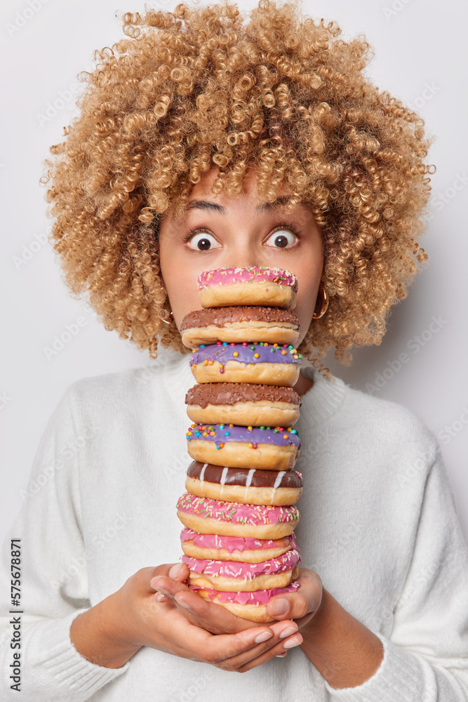 Wall mural photo of shocked curly haired woman holds stack of delicious appetizing doughnuts eats harmful food 