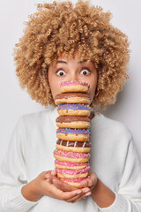 Photo of shocked curly haired woman holds stack of delicious appetizing doughnuts eats harmful food...
