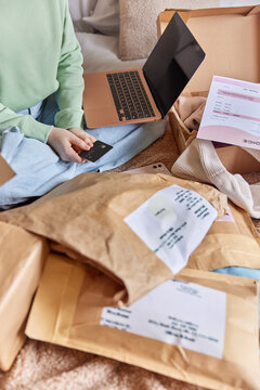Cropped image of unrecognizable faceless woman uses credit card and laptop for payment online surrounded by paper purchases going to unpack received packages poses on bed. Delivery service concept