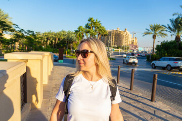 Dubai resort area, a blonde tourist girl admires the stunning view of modern architece, bustling harbor, opulent boats, towering skyscrapers, and a bright sun with clouds while taking some selfies.