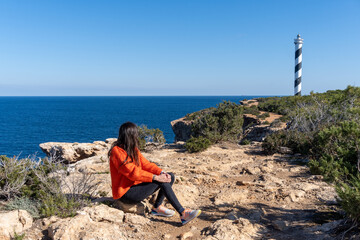 Girl looking at the Portinatx lighthouse, wearing an orange sweater, on a sunny day.