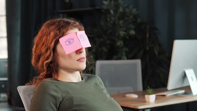 Closeup sleeping girl, funny lazy caucasian business woman tired, yawns. Sleepy female worker with sticky notes on eyes glasses with stickers sleeps in office feeling fatigued napping at workplace.