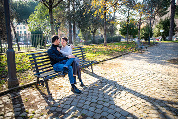 The young couple is sitting on a bench in the park in Rome. The beautiful couple in love is looking in the eyes - 575694860