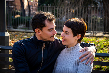 The young couple is sitting on a bench in the park in Rome. The beautiful couple in love is looking in the eyes - 575695252