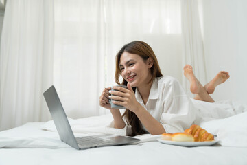 Obraz na płótnie Canvas young asian woman using laptop when sitting on a bed. Smart living with communication technology for a better life. girl sitting on at white bedroom space with a clean interior design.