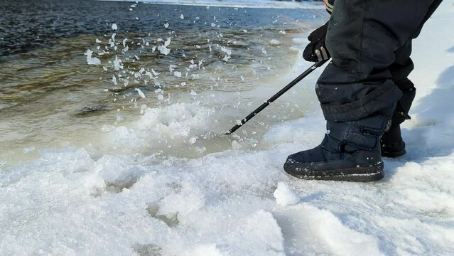 A child with a stick hammers the melted ice and snow on the river. Legs and arms close-up.