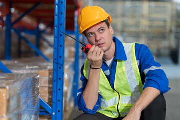 Male warehouse worker wearing hard hat and uniform using radio communication in the storage...