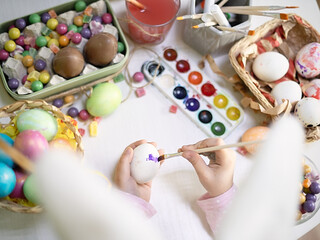 Flatlay of colorful easter eggs with candies on white table, child wearing bunny ears, kid's hands painting eggs. Overhead soft focus.