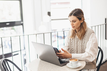 Woman holding smartphone and using laptop on table and coffee in cafe on windows background. Girl using smart phone for business, online shopping, transfer money, financial, internet banking.