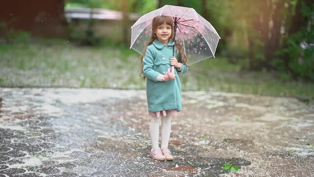 A beautiful little girl with brown hair and a toothless smile, wearing a coat under the umbrella during the falling white petals. Spring footage. Soft focus. 