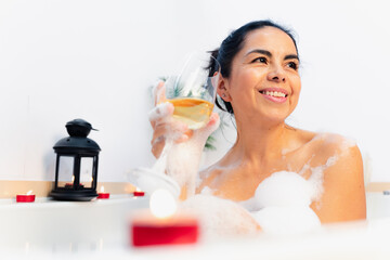 Pretty latin woman smiling while taking a bath and having a glass of wine from the bathtub of home. 