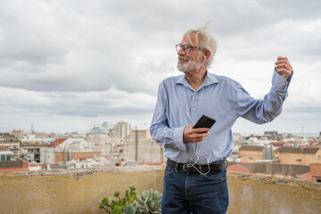 Mature businessman with gray hair and beard in blue shirt, jeans and glasses listen to music with earphones and dancing on rooftop terrace with city skylight on background. Copy space