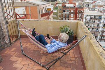 Mature business man with gray hair and beard in blue shirt and jeans laying in hammock with smartphone on terrace