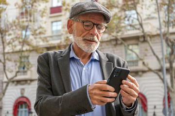 Mature man with gray hair and beard in blue shirt, jacket and cap standing on a street with buildings and texting with smartphone
