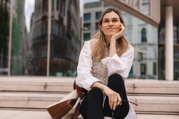 Obraz premium Attractive blonde woman sitting on stairs near office building and look happy. Blonde girl smiling and look at side. Amazed woman wear beige knit vest, white shirt.
