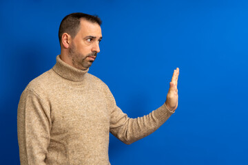 Strict bearded hispanic man showing stop sign, standing with frowning and bossy expression, meaning caution to avoid danger or mistake. Isolated on blue background