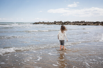 rear view of little girl with blown hair standing amidst waves on sand