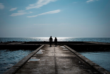 A couple walking towards the sea on a bridge on a beautiful evening to wave the sun and a small yacht in between them
